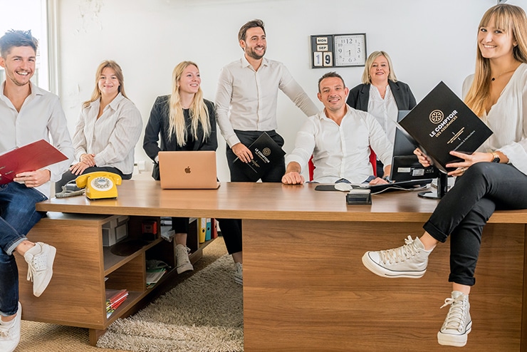 Équipe Le Comptoir des Assurances souriante au bureau Sept souriants professionnels, vêtus de blanc et noir, posent autour d'un grand bureau avec un téléphone jaune et un cahier "LE COMPTOIR DES ASSURANCES".