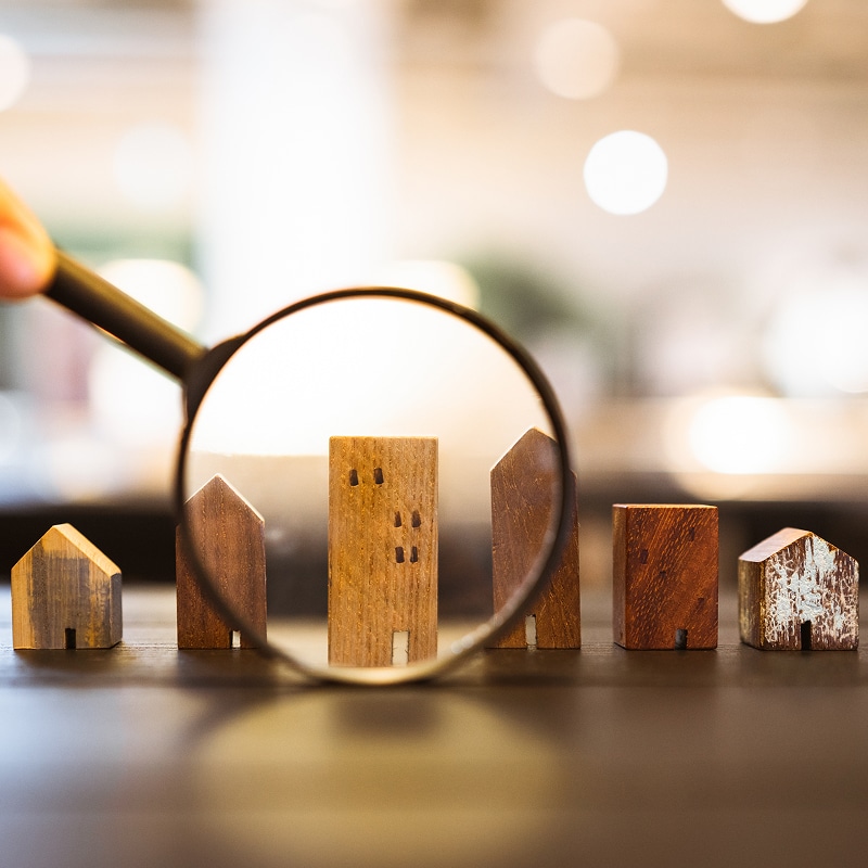 Loupe examinant un immeuble en bois miniature, parmi d'autres maisons, illustrant la recherche immobilière ou l'inspection de propriété.