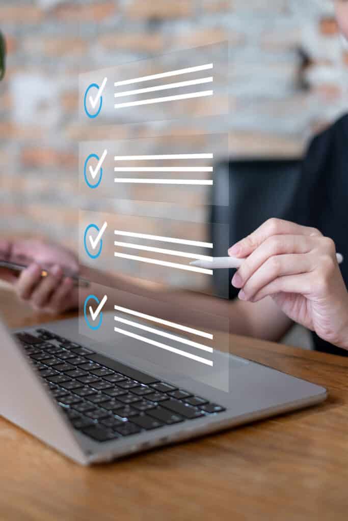Gestion de Tâches Numériques : Cocher une Checklist Main utilisant un stylet pour cocher des éléments sur une liste de tâches virtuelle flottante au-dessus d'un ordinateur portable.