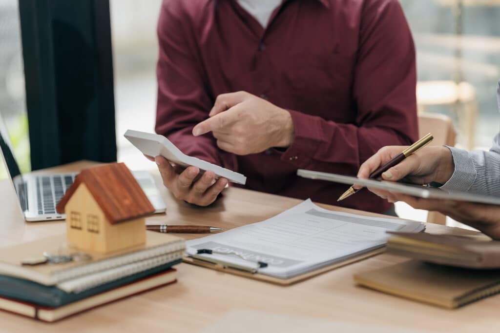 Signature de contrat immobilier : calcul et négociation Deux personnes discutent de chiffres sur une calculatrice et un contrat, près d'une maquette de maison en bois.