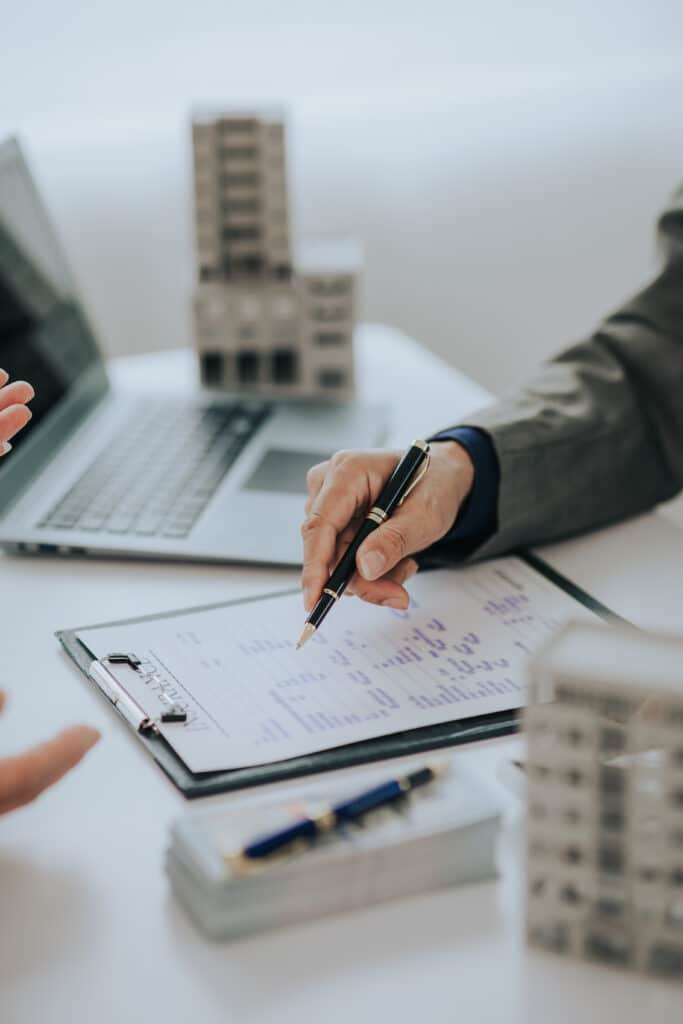 Signature Contrat Assurance : Affaires et Immobilier Main en costume pointant avec un stylo noir et or un document 'INSURANCE' sur un presse-papiers, près d'un ordinateur portable et de modèles de bâtiments.