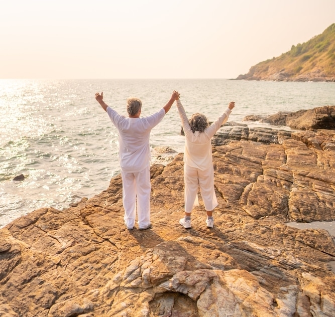 Couple senior: la liberté retrouvée au bord de la mer. Couple senior main dans la main sur la côte rocheuse, les bras levés vers l'océan au coucher du soleil.