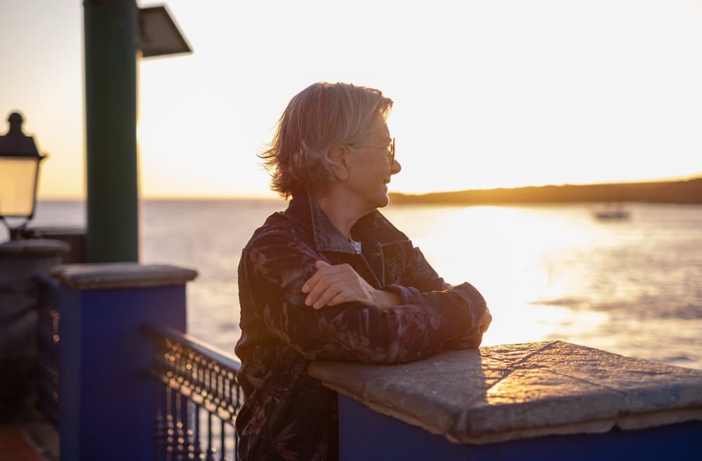 Coucher de soleil : Femme âgée contemplative en bord de mer. Femme âgée souriante, cheveux gris, appuyée sur une balustrade en bord de mer. Contemplation au coucher du soleil sur l'océan.